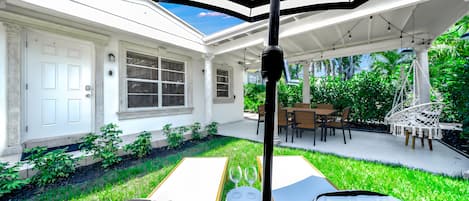 Un patio cubierto con una mesa y sillas para comer, rodeado de abundante follaje y un edificio blanco con ventanas.