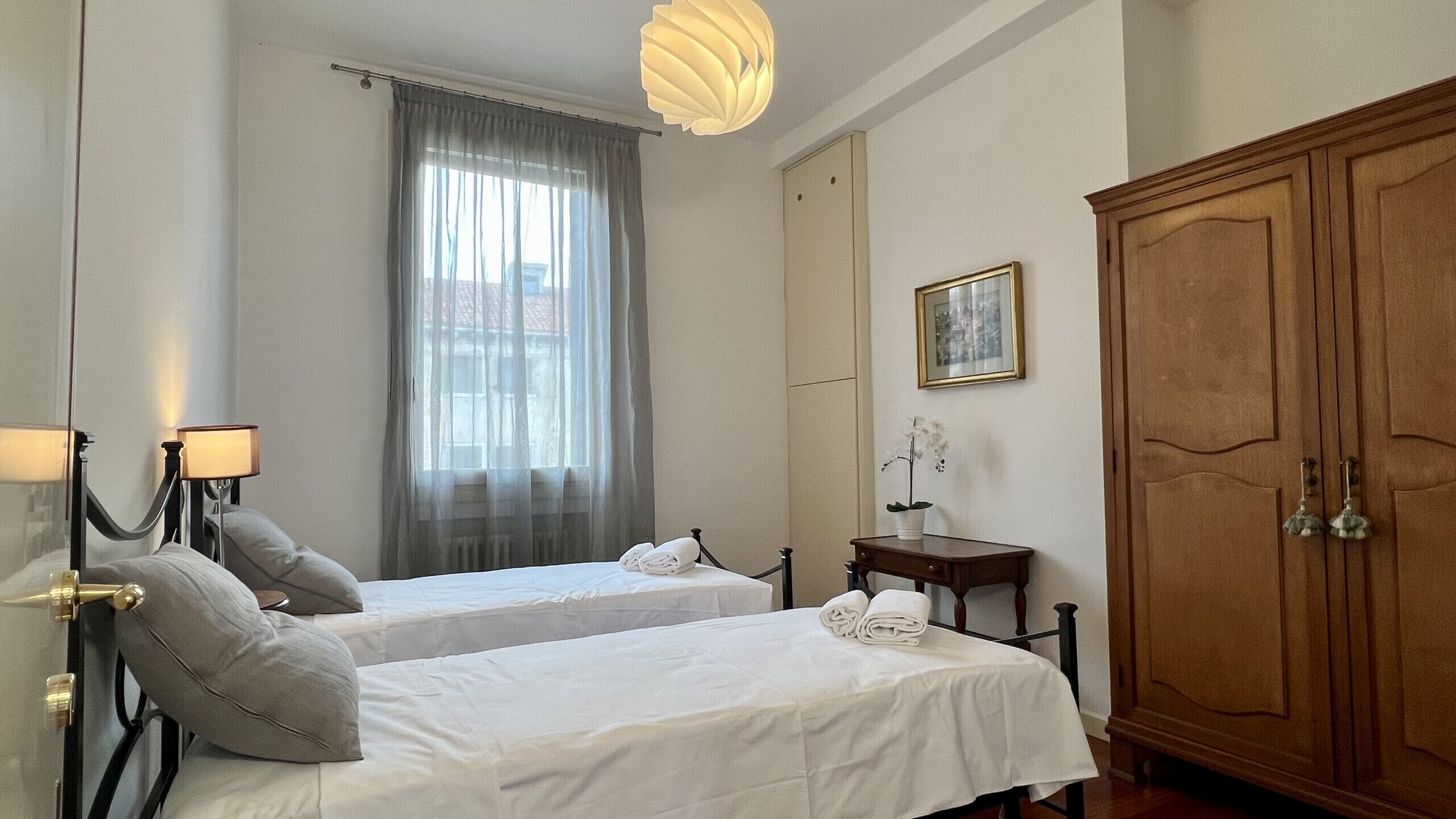 Quarto de hotel com duas camas, um guarda-roupa de madeira, uma mesa de cabeceira com abajur e uma janela com cortinas.