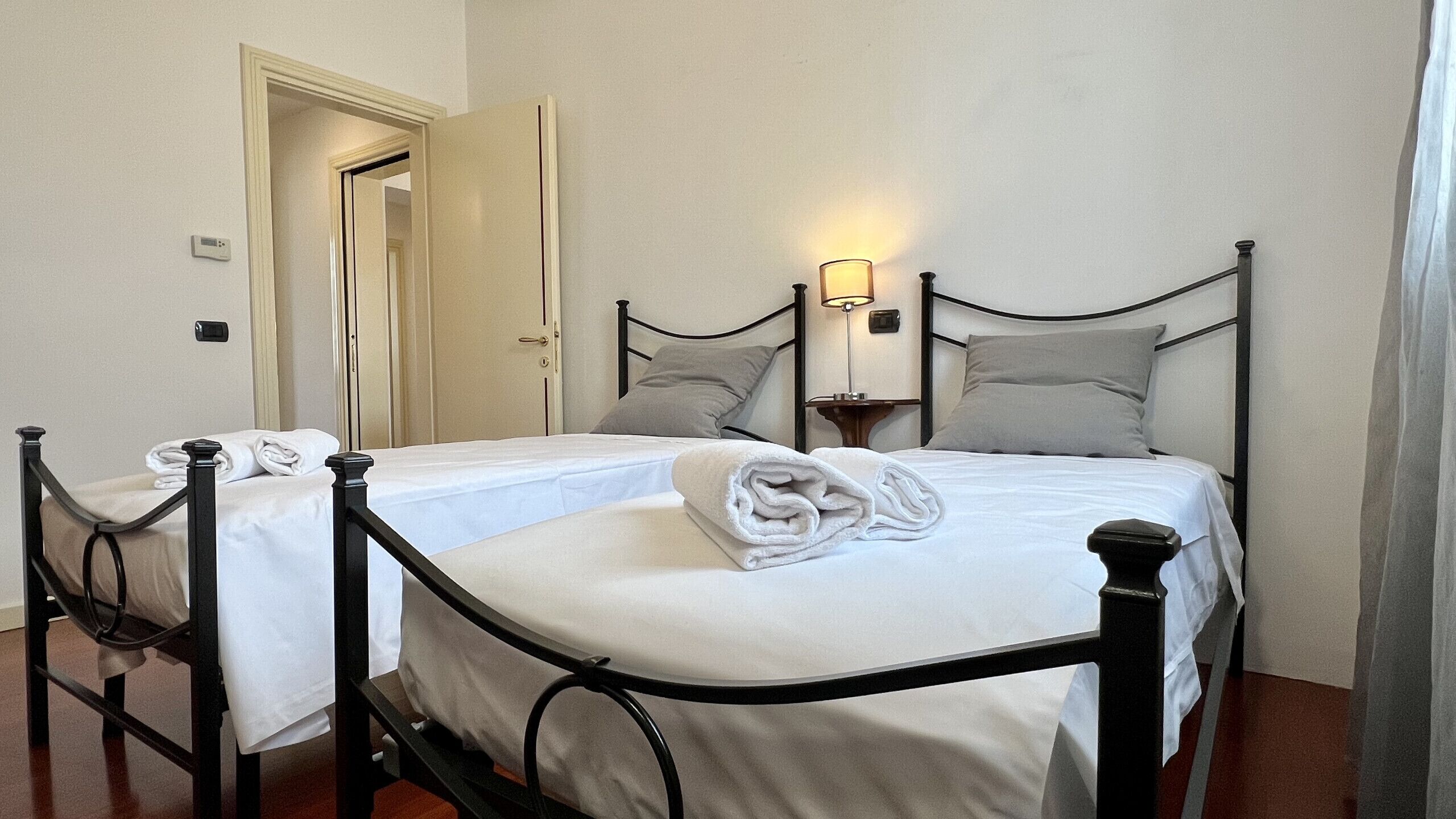 Un lit double avec un cadre en métal noir, du linge de lit blanc et deux oreillers. Il y a une table de chevet avec une lampe, une porte donnant sur une autre pièce et un thermostat au mur.