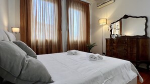 Ein ordentlich gemachtes Bett mit weißen Leinen und zwei aufgerollten Handtüchern, eine Holzkommode mit Spiegel sowie eine Stehlampe.