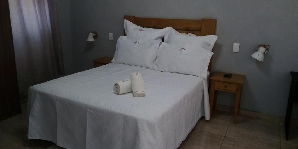 Una cama bien hecha con sábanas y almohadas blancas, un cabecero de madera, dos mesitas de noche con lámparas y una cortina parcialmente abierta.