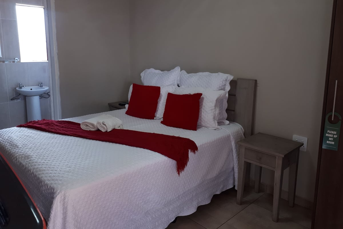 Een net opgemaakt bed met wit beddengoed en rode kussens, een klein nachtkastje en een wastafel aan de muur.