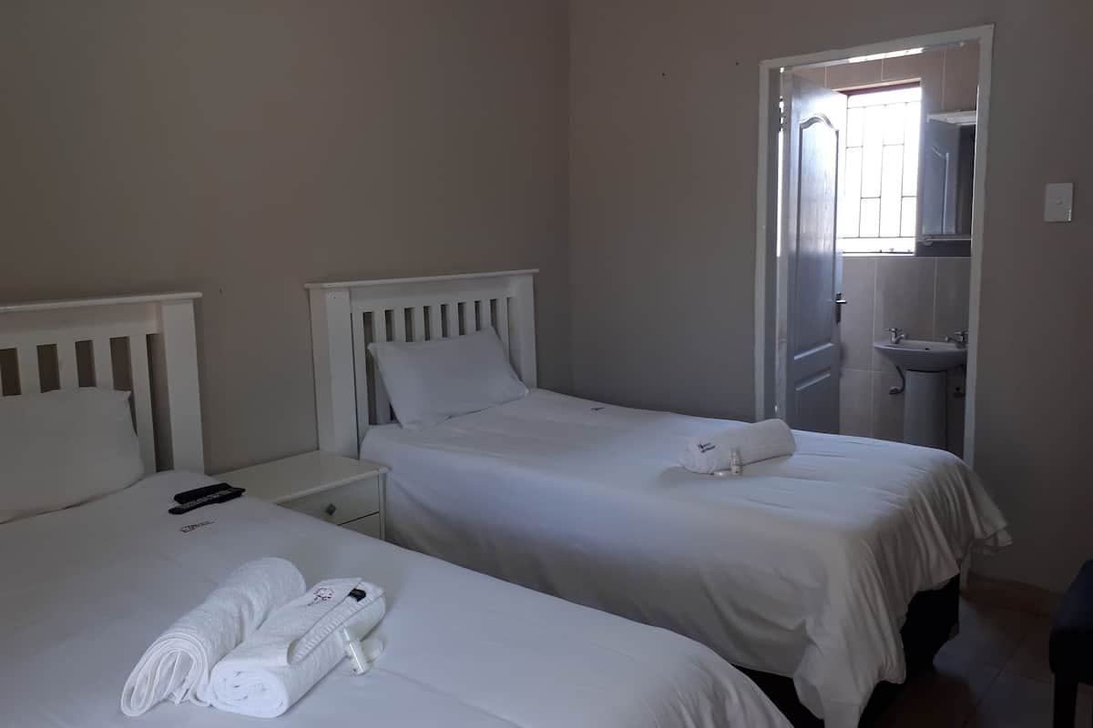 Een hotelkamer met twee enkele bedden, elk met een witte hoofdbord en passende kussens. Er staat een klein nachtkastje met een afstandsbediening en een fles lotion. Een handdoek is netjes op het bed gevouwen.
