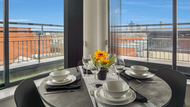 A dining table set for four with white plates, bowls, and cutlery, a vase with sunflowers, and a view of a cityscape through large windows. - Penthouse With City Skyline Views - Balcony  - King Bed - Netflix (birmingham)