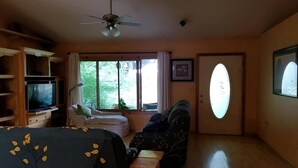 Ein Zimmer mit einem Bett, einem Fernseher, einem Deckenventilator, einem Fenster mit Vorhängen, einer Tür mit einem ovalen Spiegel und einem kleinen Tisch.
