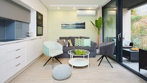 A modern kitchen with white cabinets, a built-in oven, and a microwave. There is a dining area with a round table, two chairs, and a small round ottoman. A gray sofa with patterned cushions is placed in front of a large window, and a painting hangs on the wall. - Hedera A12 (Dubrovnik)