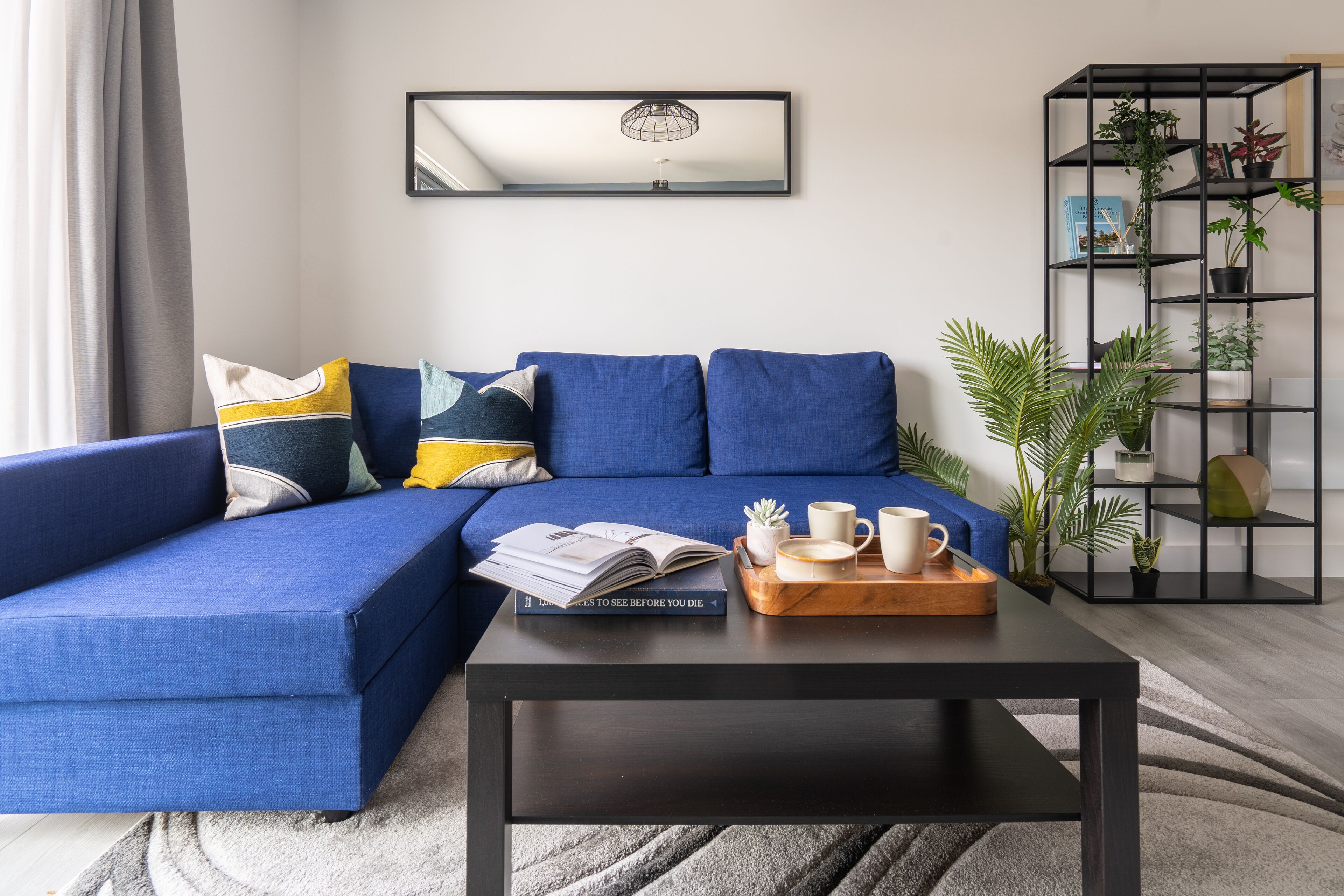 Un salon moderne avec un canapé d’angle bleu, une table basse noire et une étagère noire agrémentée de plantes.