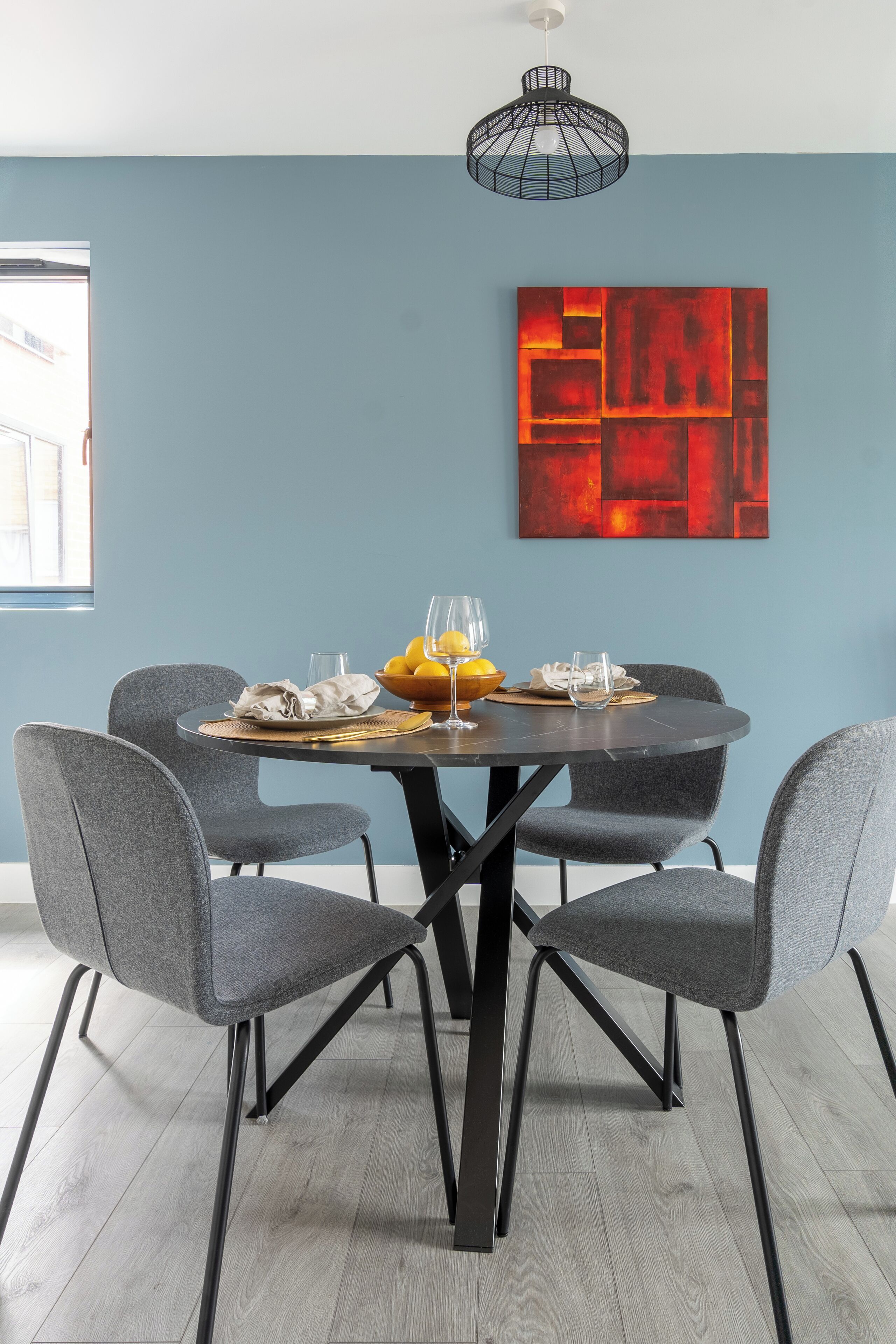 Un comedor con una mesa redonda, cuatro sillas grises, estructura de metal negro, un cuadro abstracto rojo y una lámpara colgante.