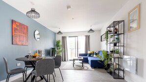 A modern living room with a blue wall, a dining area with grey chairs, a blue sofa, and a black shelving unit with plants. - Stunning 1-bed Apartment in Hemel Hempstead (Hemel Hempstead)