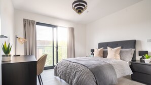 A modern bedroom with a large bed, a desk, and a chair. There is a window with curtains, a ceiling light, and a small plant on the desk. - Stunning 1-bed Apartment in Hemel Hempstead (Hemel Hempstead)