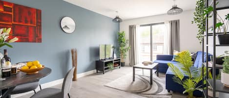 A modern living room with a blue accent wall, a dining area with a round table and chairs, a blue sofa, and a bookshelf with plants.