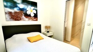 En pænt redt seng med hvidt sengetøj og en gul pude, et lille natbord med en lampe, og et billede af blomster og vand hængt på væggen.