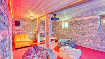 A cozy room with stone walls, a wooden ceiling, and a red carpet. There are bean bags and a bright orange sofa.