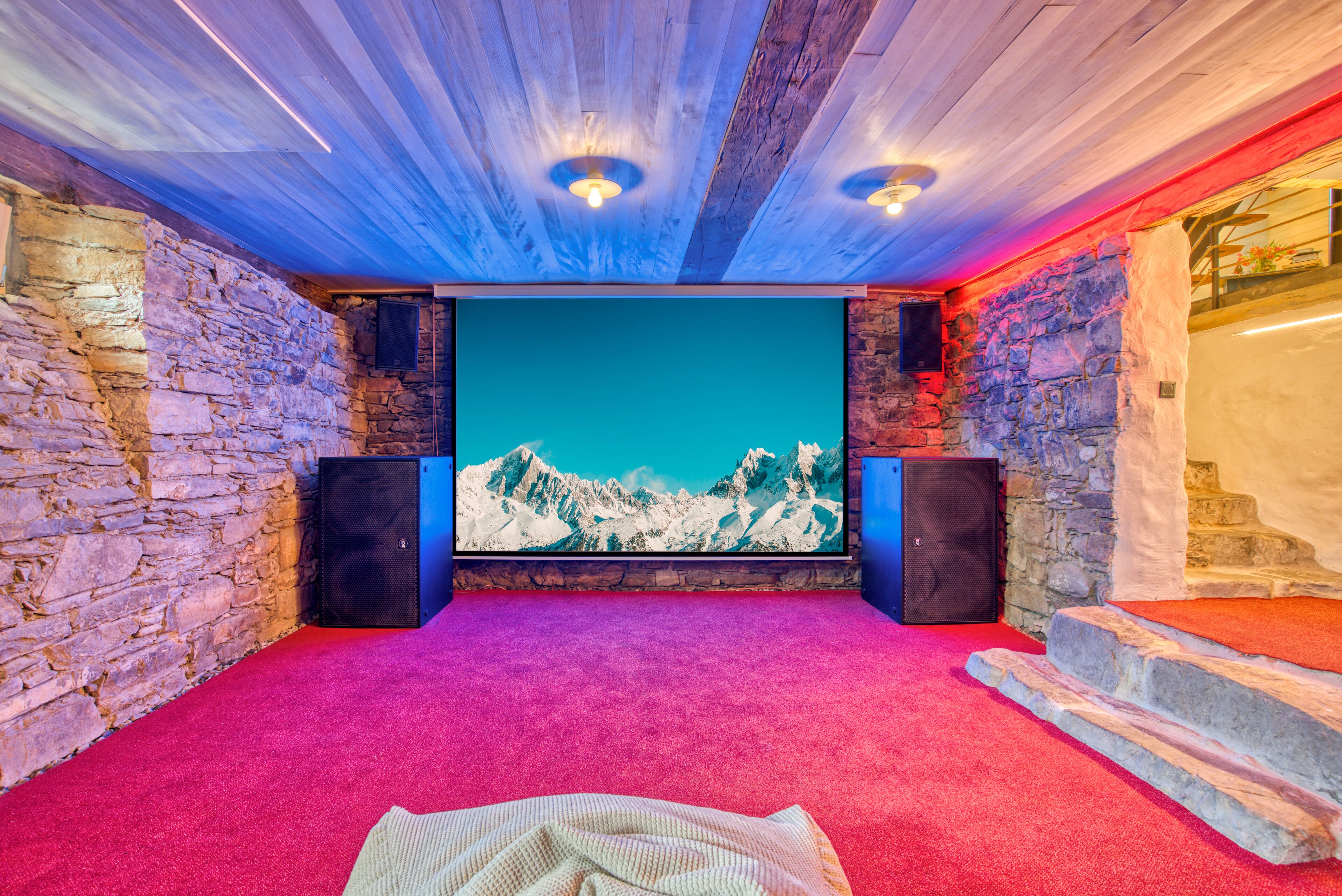 A room with a red carpet, stone walls, a wooden ceiling, and a large screen displaying a mountain scene.