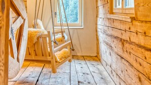 Interior rústico de una cabaña de madera con una cama en literas, un columpio colgante y una ventana que da a un paisaje de árboles.