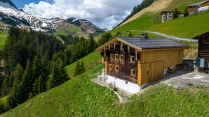 Una cabaña de madera con techo inclinado, rodeada de vegetación exuberante y montañas al fondo.