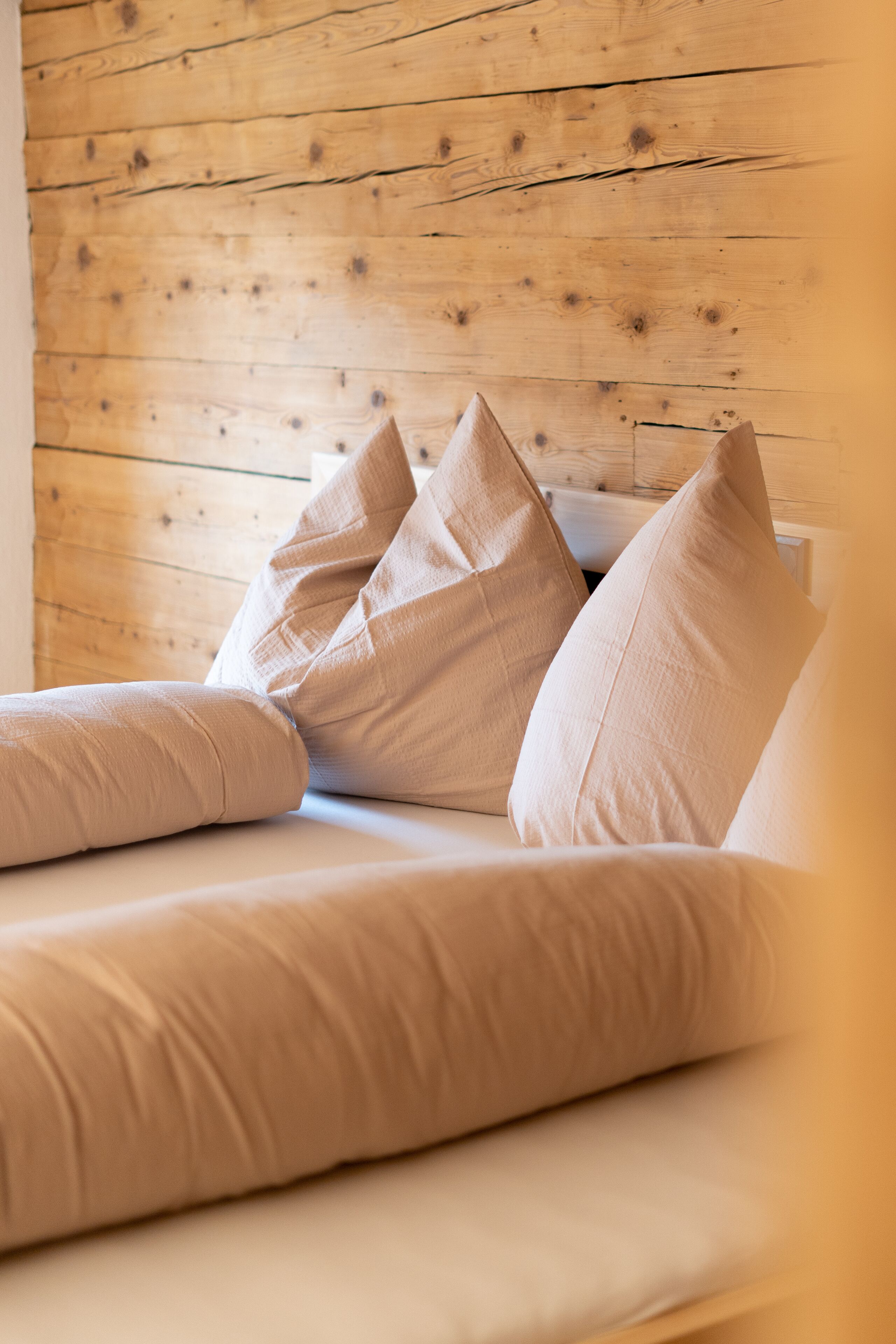 A bed with beige bedding and pillows against a wooden wall.