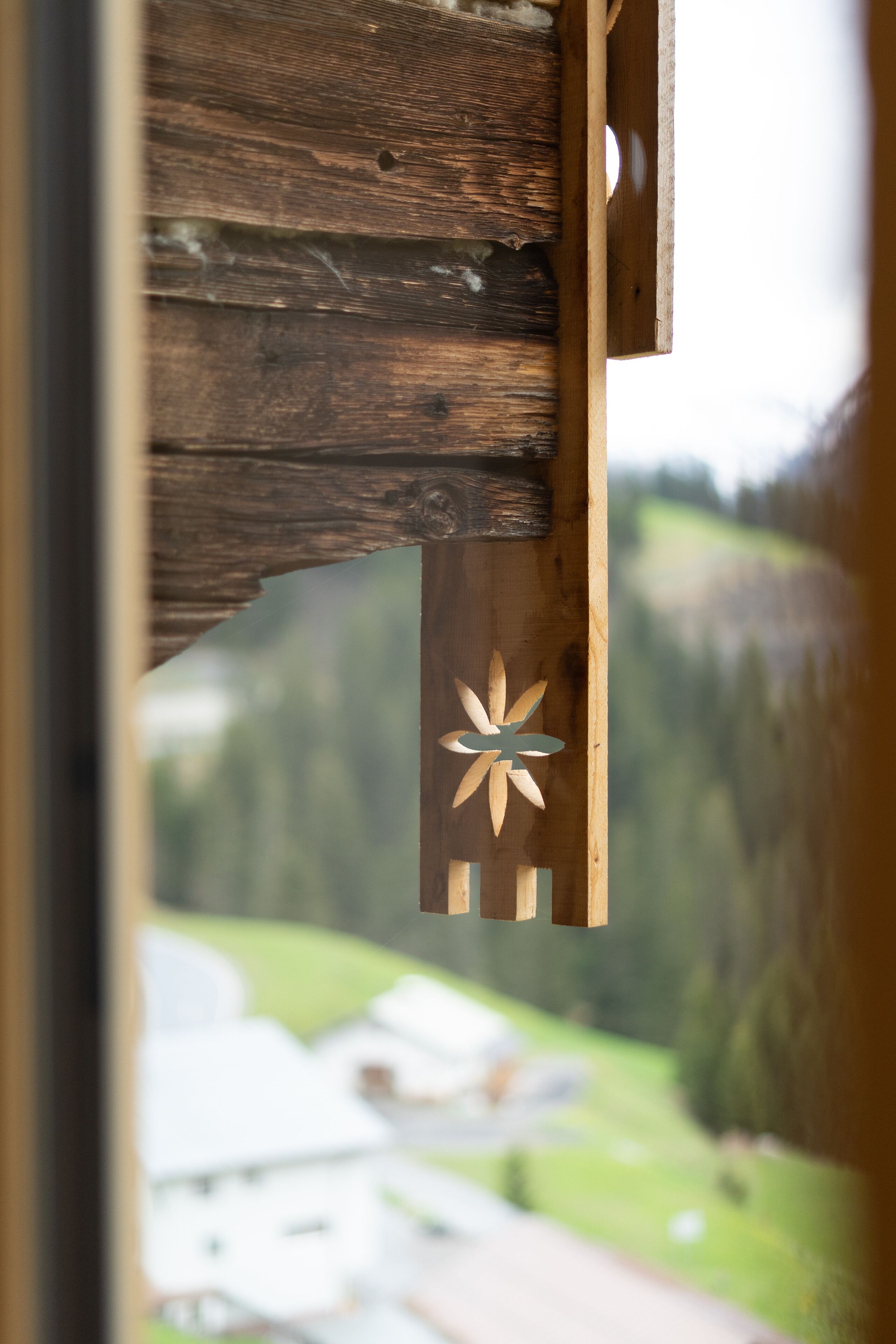 A wooden structure with a cut-out design, likely part of a window frame or door, with a blurred view of a green landscape outside.