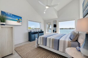 Un dormitorio con cama, ventilador de techo, ventana grande con vista al mar y un cuadro de una playa.