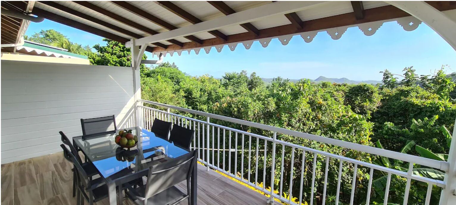 A balcony with a glass table and chairs, surrounded by a white railing, overlooking a lush green landscape with mountains in the distance.