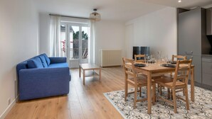 Una sala de estar moderna con un sofá azul, una mesa de comedor de madera y sillas. Se observa un ventanal con cortinas, un radiador y un aplique de techo.