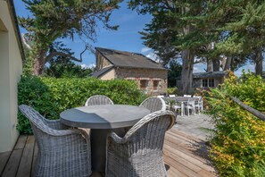 A wooden deck with wicker furniture, a stone building in the background, and surrounding greenery.