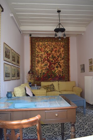 Une pièce avec un canapé à carreaux, une chaise en bois, une table avec un plateau en verre et une tapisserie représentant un arbre accrochée au mur.