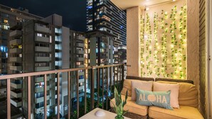 A balcony with a view of adjacent buildings, a sofa with cushions, a small table with a plant, and a wall decorated with string lights and greenery. - Renovated Waikiki Studio! City View, Full Kitchen, Free Wi-Fi & Parking, Pool (Honolulu)