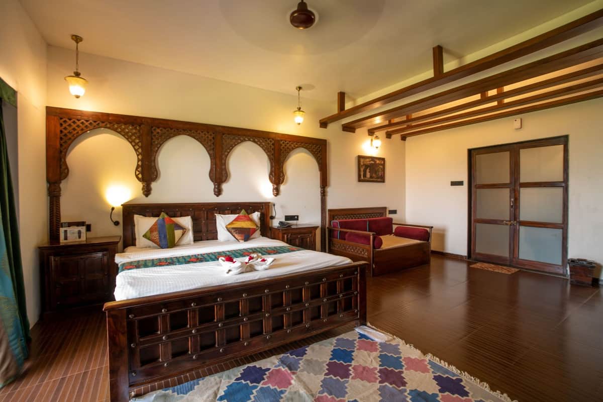 A spacious bedroom with a large bed, wooden headboard, and a decorative archway. There is a small seating area with a red chair and a wooden bench. The room has a wooden floor and a large window with a view of the outside.