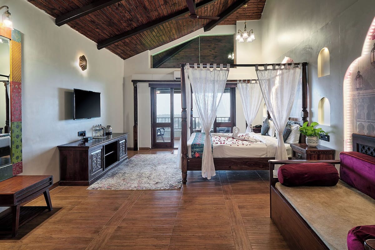 A bedroom with a four-poster bed, a television, a wooden floor, and a balcony with a view.