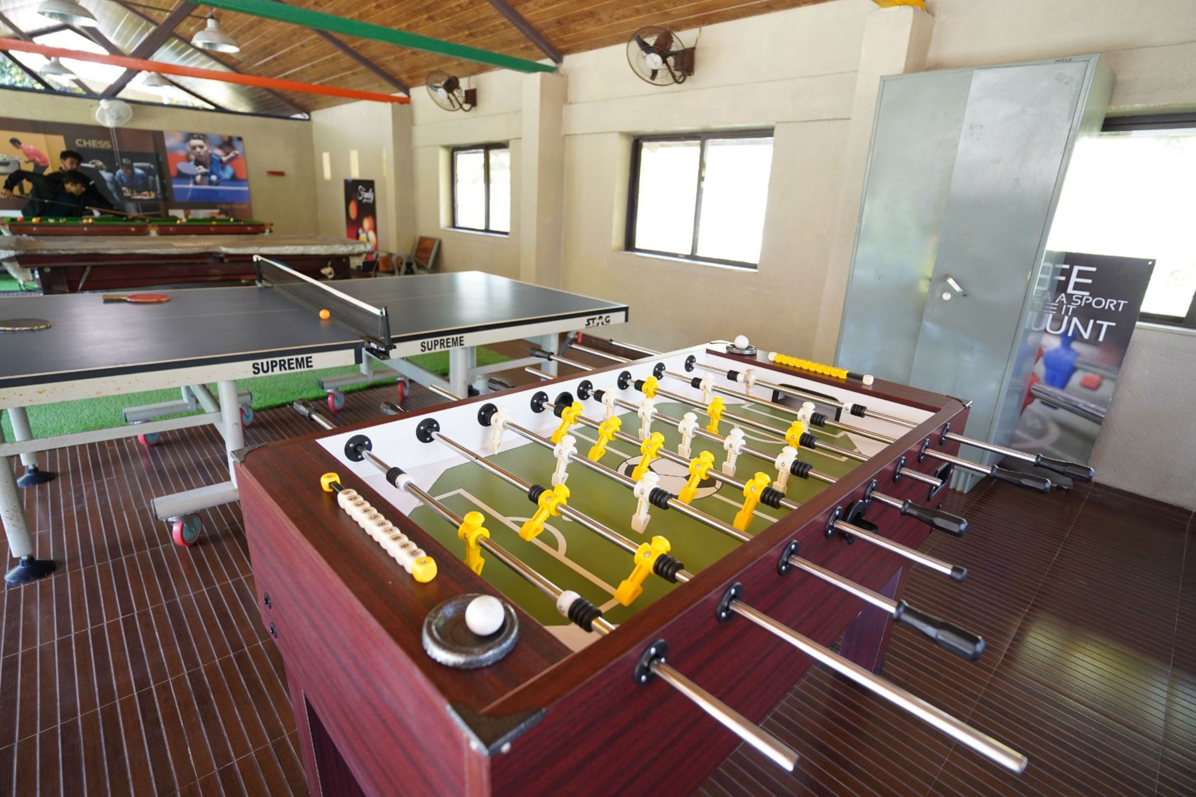 Una habitación con mesa de fútbol, mesa de ping pong y mesa de ajedrez. Hay ventanas y un ventilador en la pared.