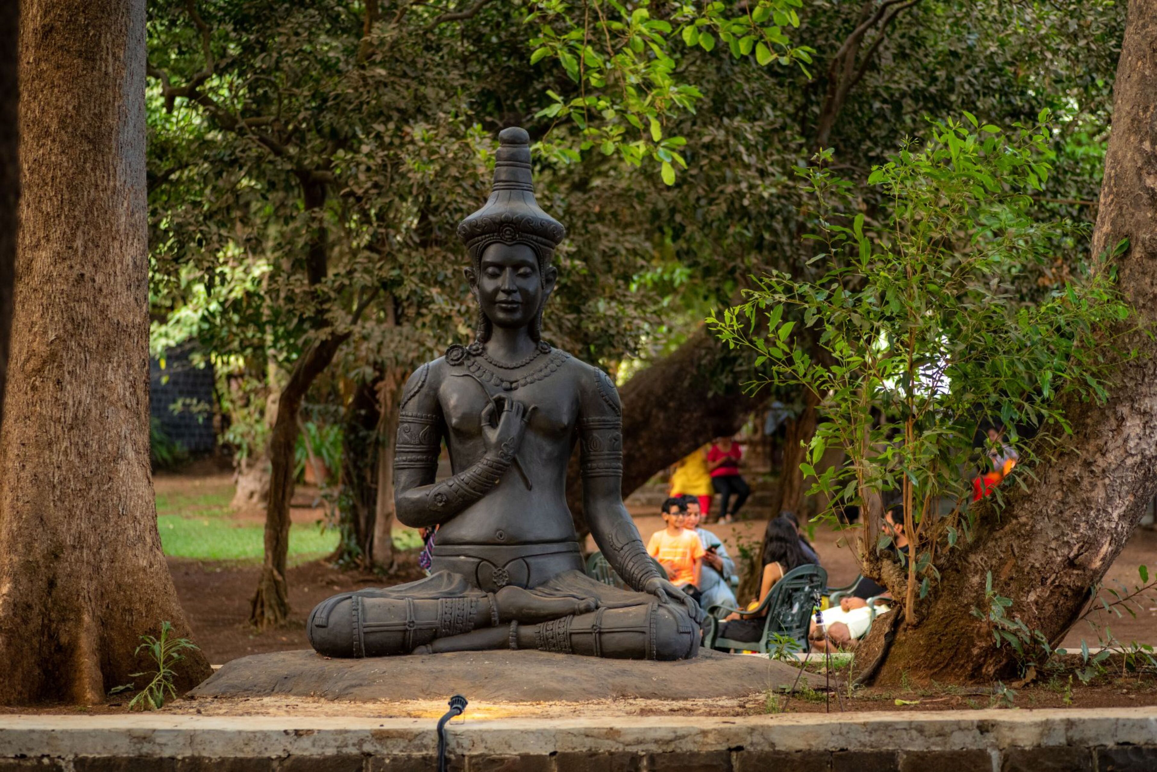 A seated statue in a park with trees and people in the background.