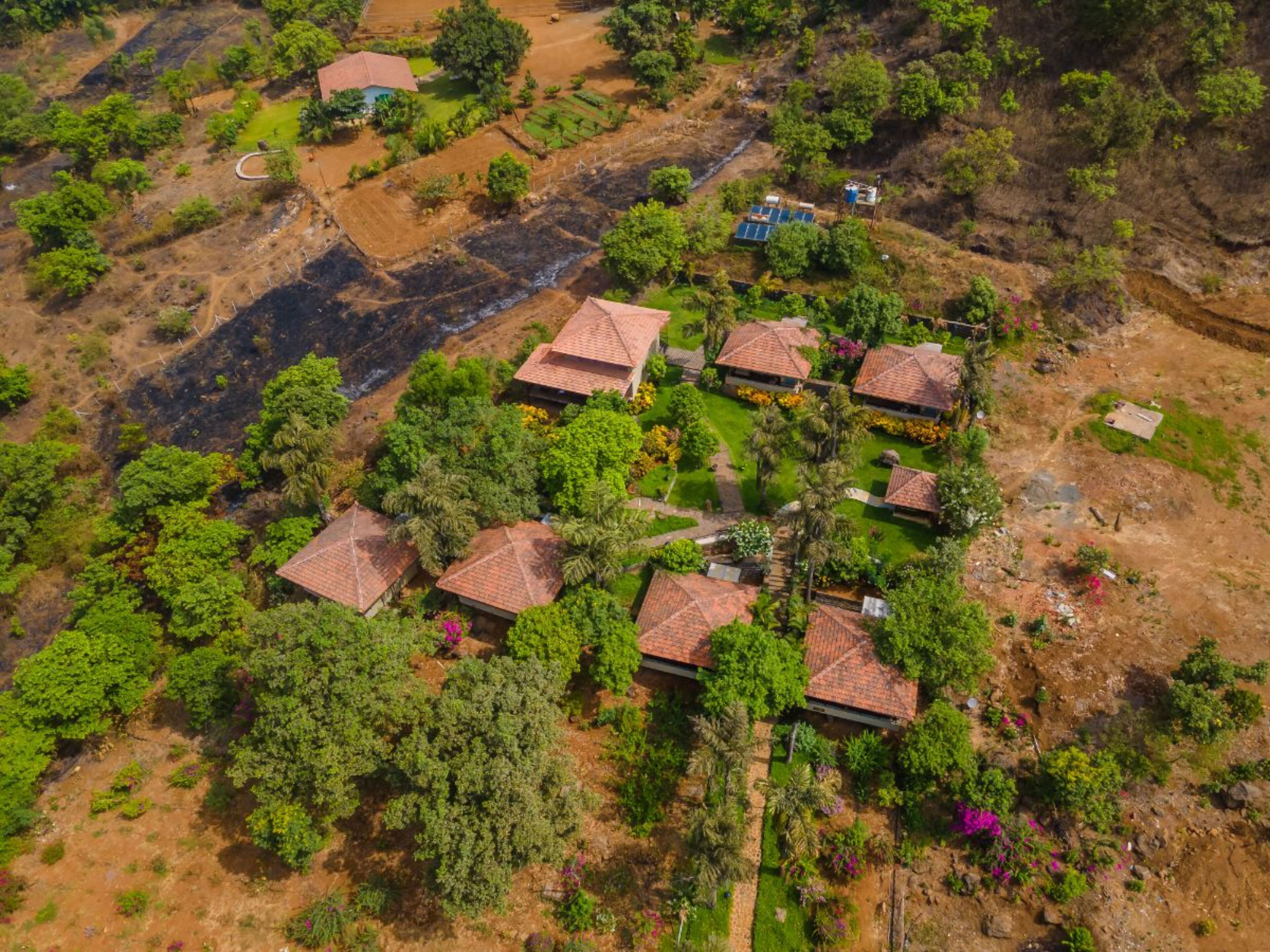An aerial view of a rural village with several houses featuring red-tiled roofs, surrounded by lush greenery and scattered trees.