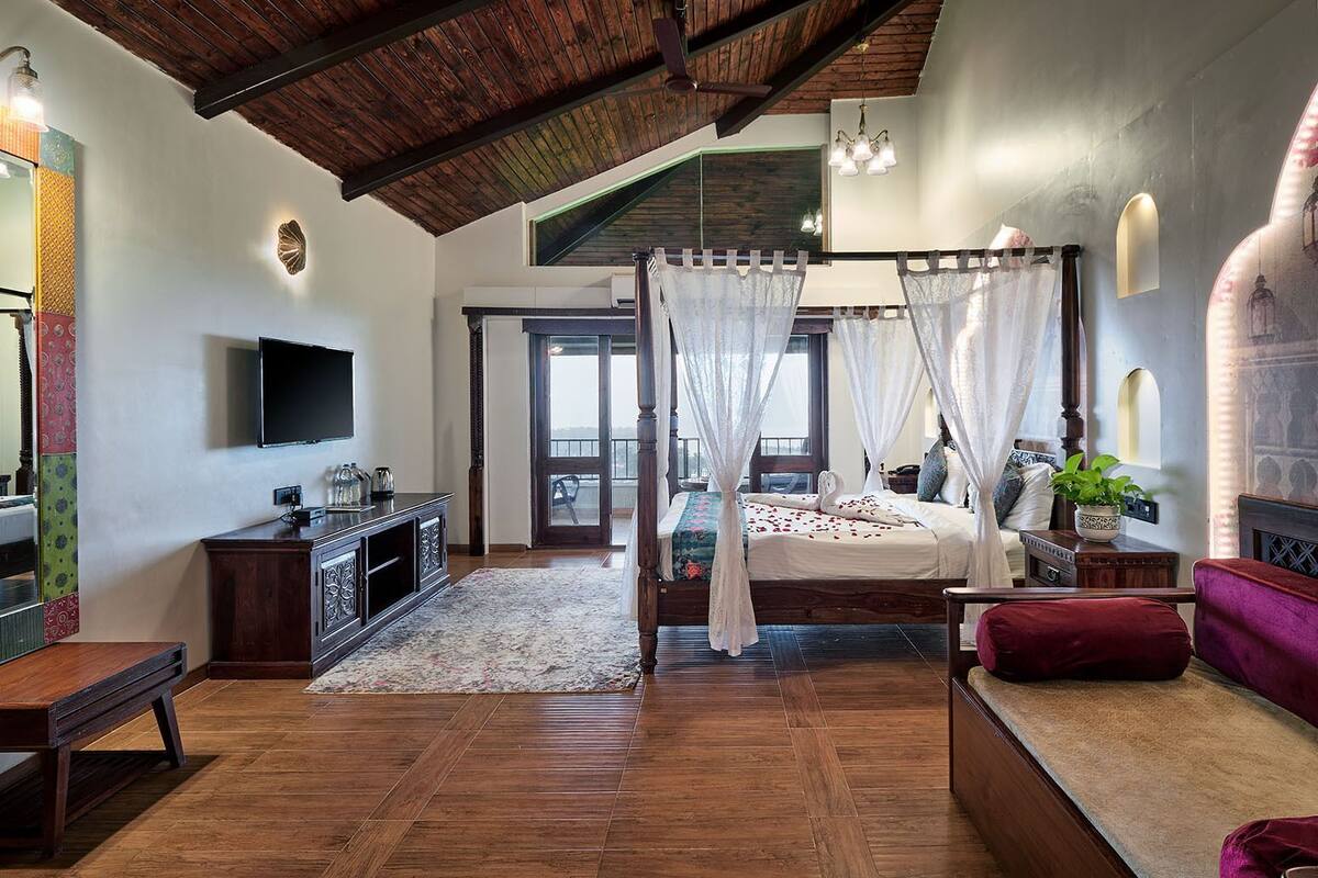 A bedroom with a four-poster bed, a TV, a wooden bench, and a view of the outside.