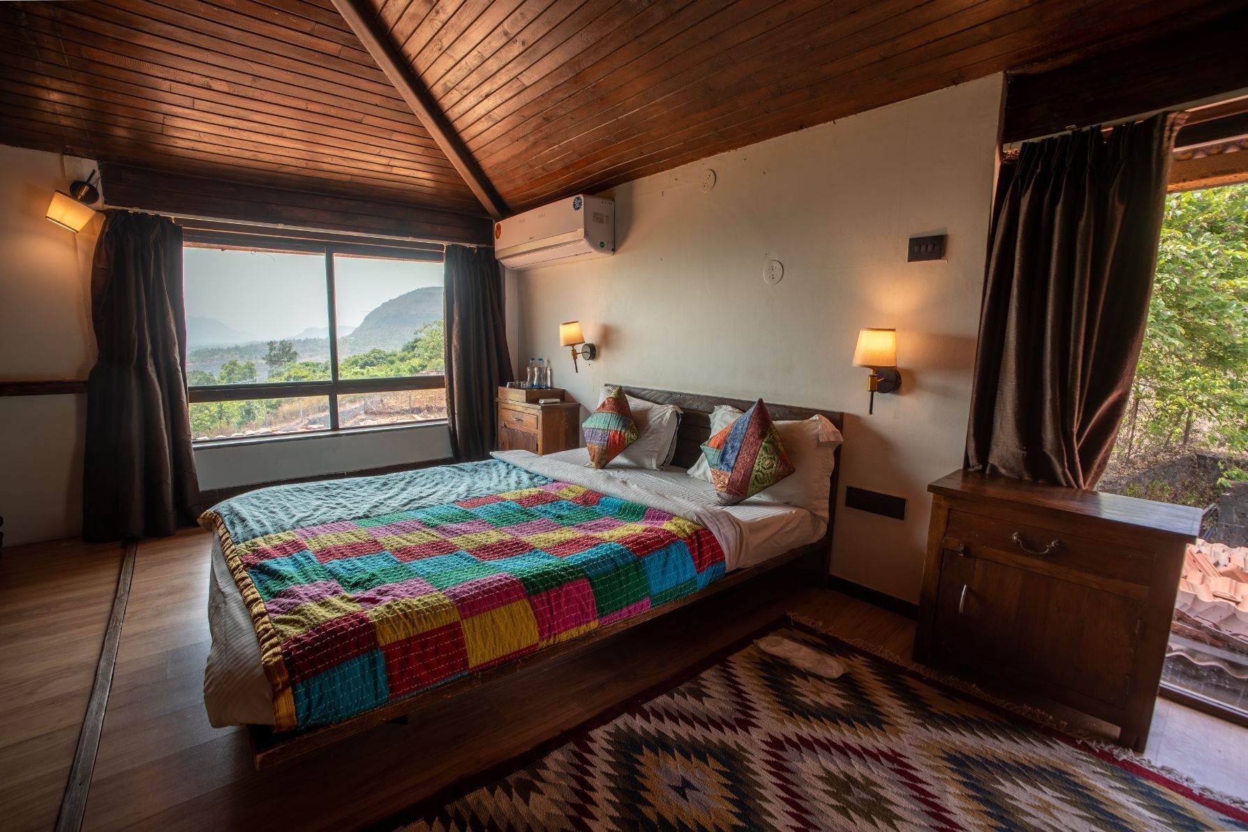 Ein Schlafzimmer mit einem großen Bett, Holzboden, einem Fenster mit Vorhängen, einem Nachttisch und einer Wandlampe.