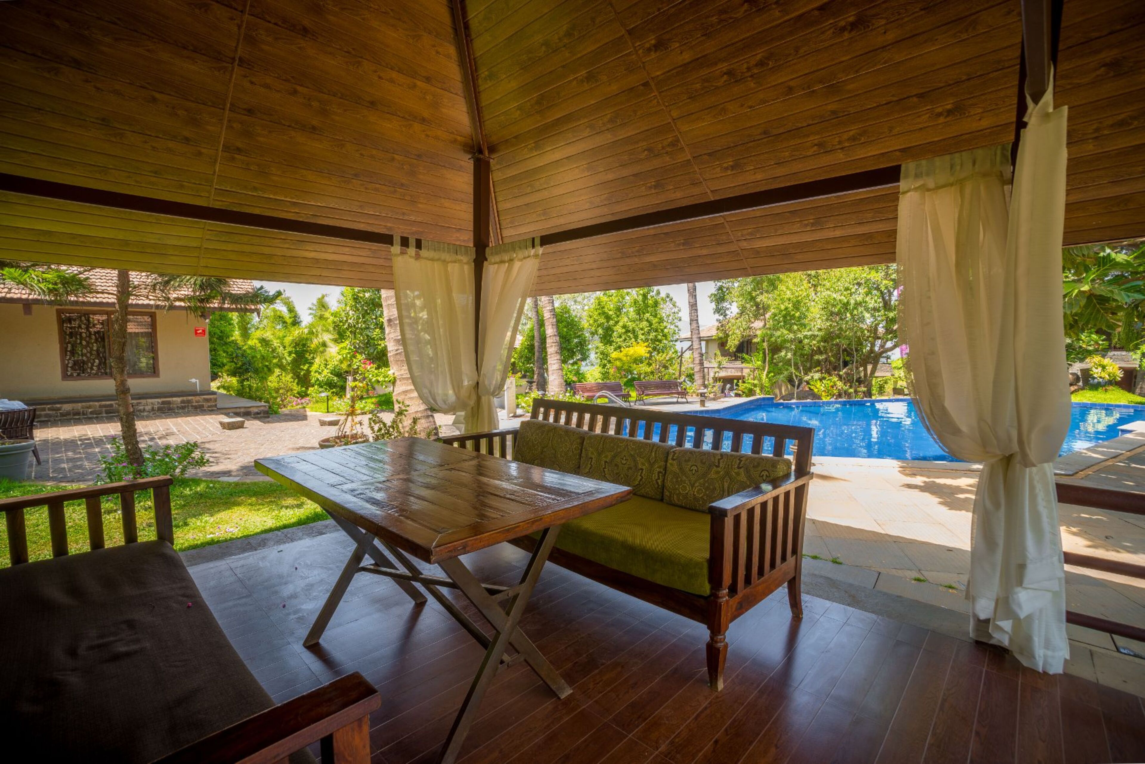 An outdoor patio with a wooden table and bench, a pool in the background, and a view of the surrounding greenery.