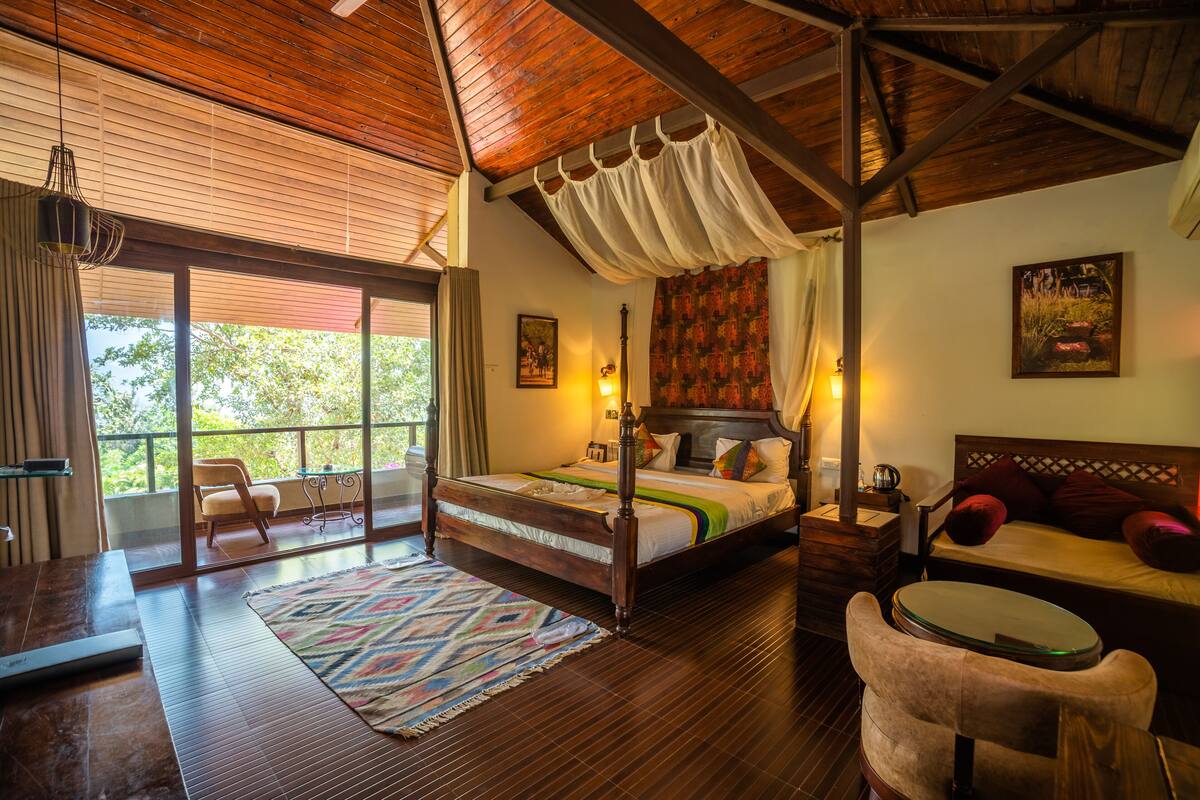A spacious bedroom with a large bed, a sitting area with a table, and a view of greenery through the windows.