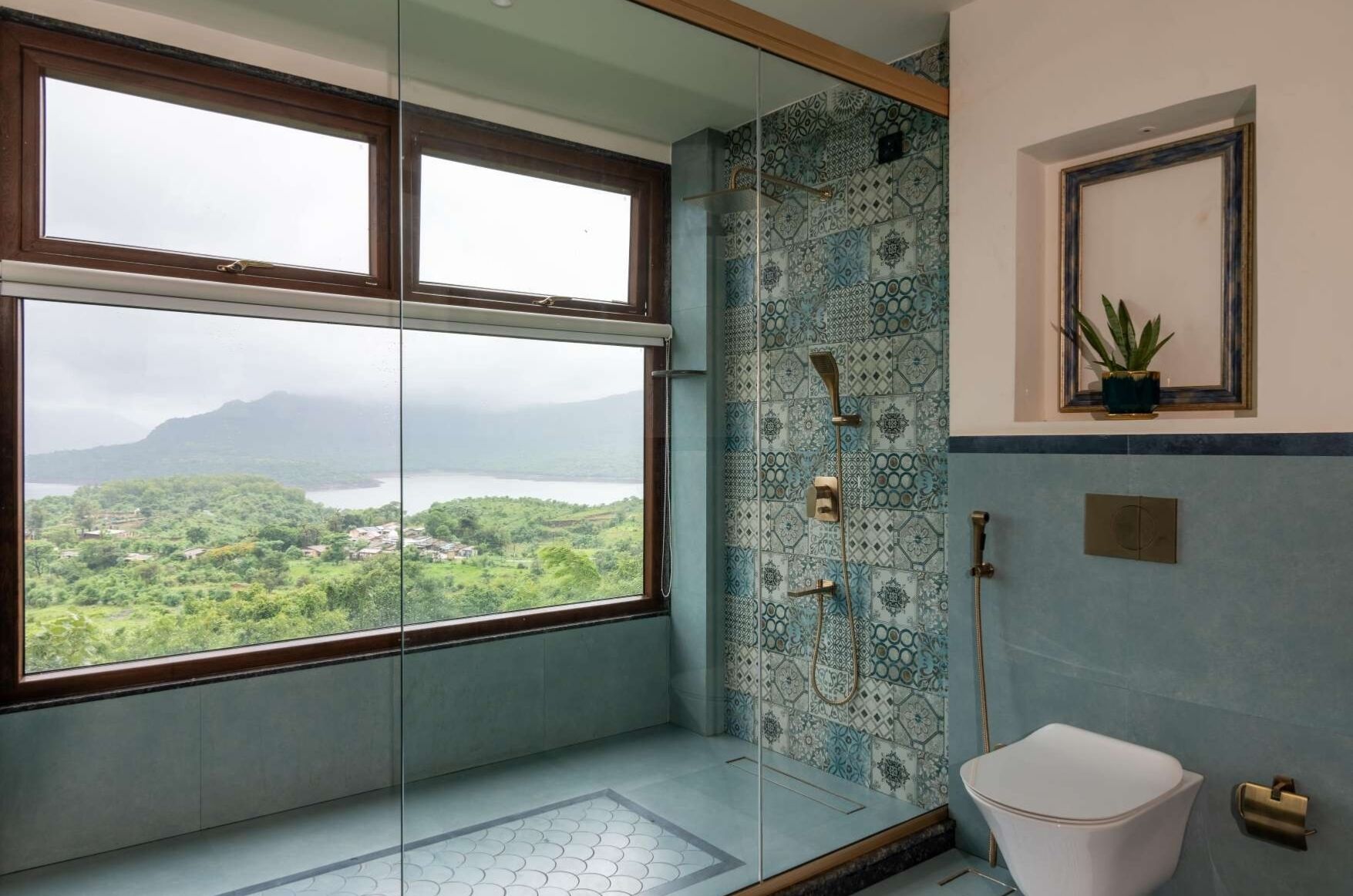 Un baño con una ventana grande, una ducha con pared de azulejos decorativos, un inodoro y una pequeña planta en maceta.