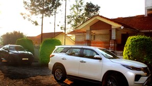 A white SUV parked in front of a house with a tiled roof and a brick wall. - Yercaud Bungalow Stay (Yercaud)
