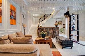 Amplia sala de estar con un sofá beige grande, una mesa de centro de madera y un reposapiés negro. Una escalera conduce a un nivel superior, y se ve una estantería con objetos decorativos a la derecha.