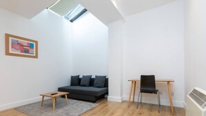 Una sala de estar moderna con un sofá gris oscuro, una mesita de madera, una silla negra y una alfombra gris. Hay un tragaluz sobre el sofá y un radiador blanco a la derecha.