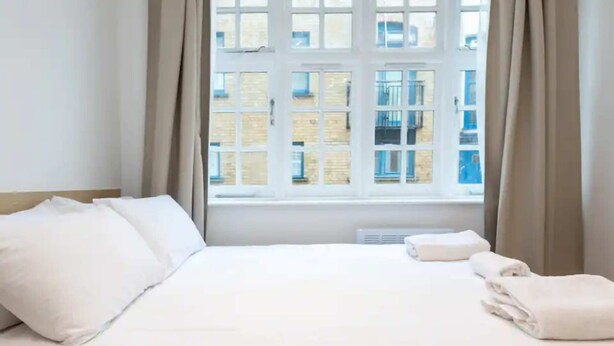 Una cama bien hecha con sábanas y almohadas blancas, una ventana con cortinas y vistas a edificios.