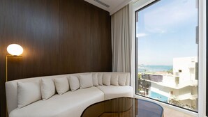Una sala de estar moderna con un sofá curvo, una mesa de centro de vidrio y un ventanal con vistas al mar y edificios.