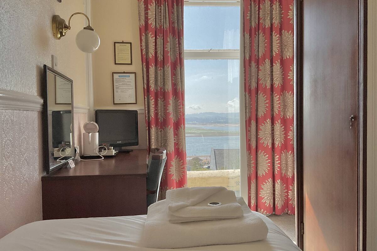 Ett hotellrum med en säng, ett skrivbord med en tv, en lampa och utsikt över havet genom ett fönster med blommiga gardiner.
