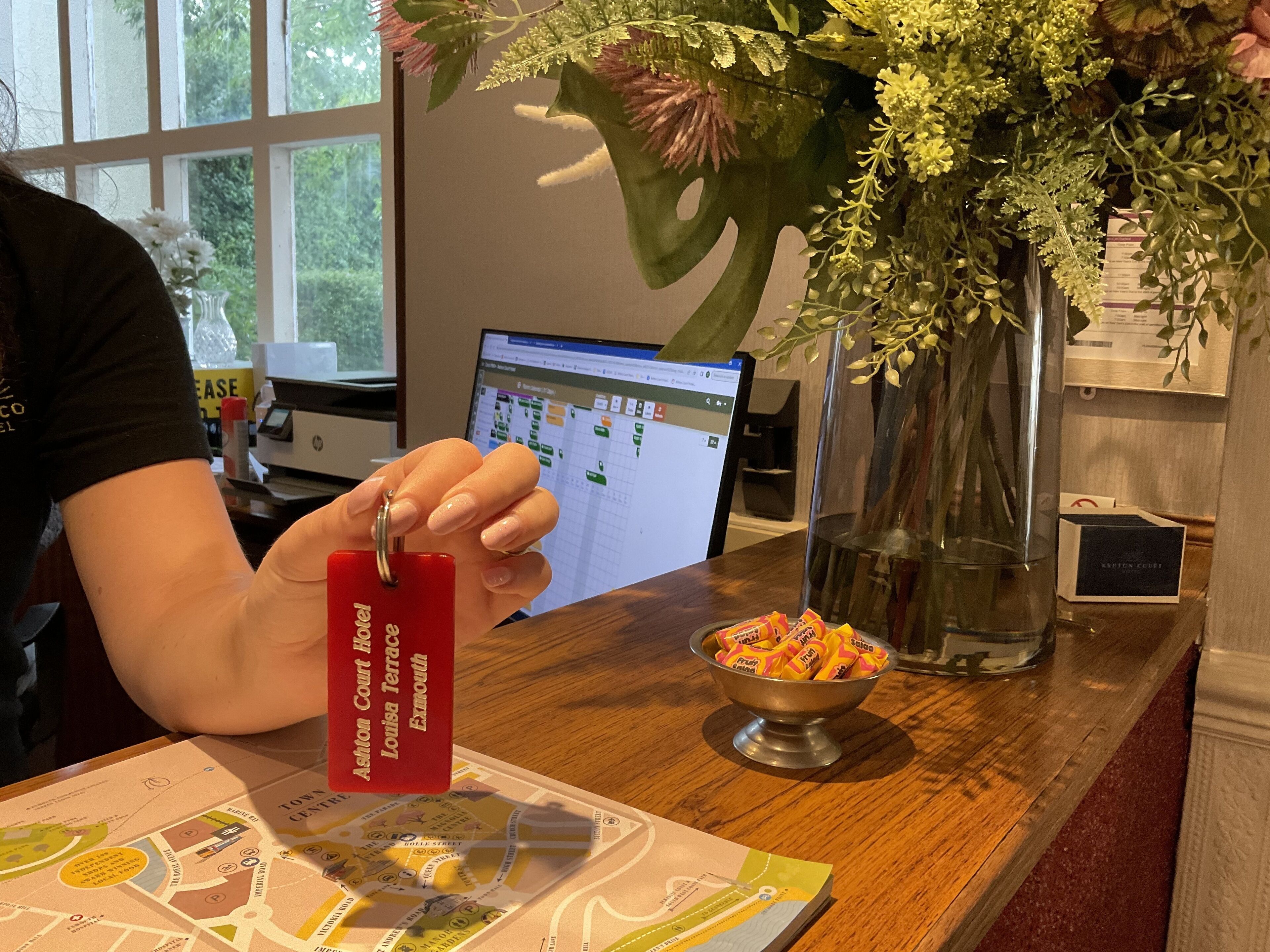 A person holding a keychain with hotel names, a bowl of fruit, a vase with flowers, and a map on a wooden table.