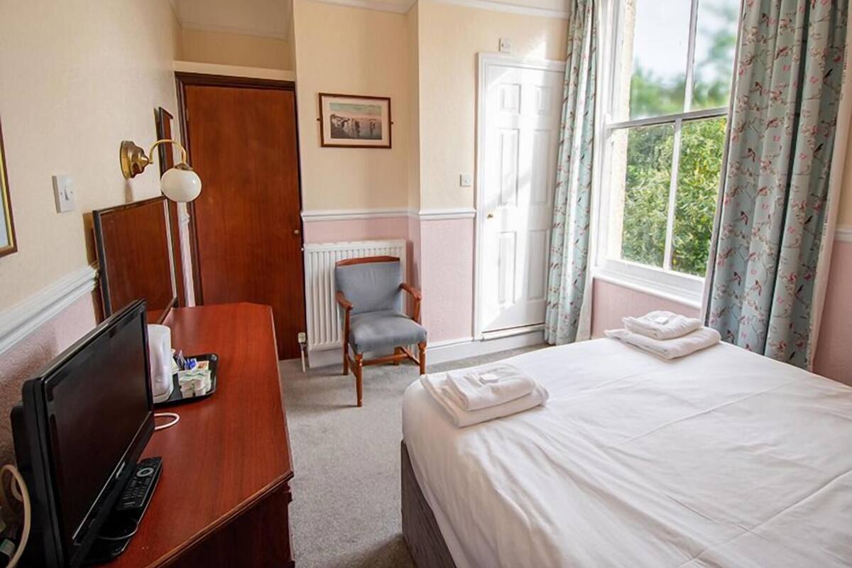 Ett hotellrum med en säng, ett skrivbord med en dator, en stol, ett element och ett fönster med gardiner.
