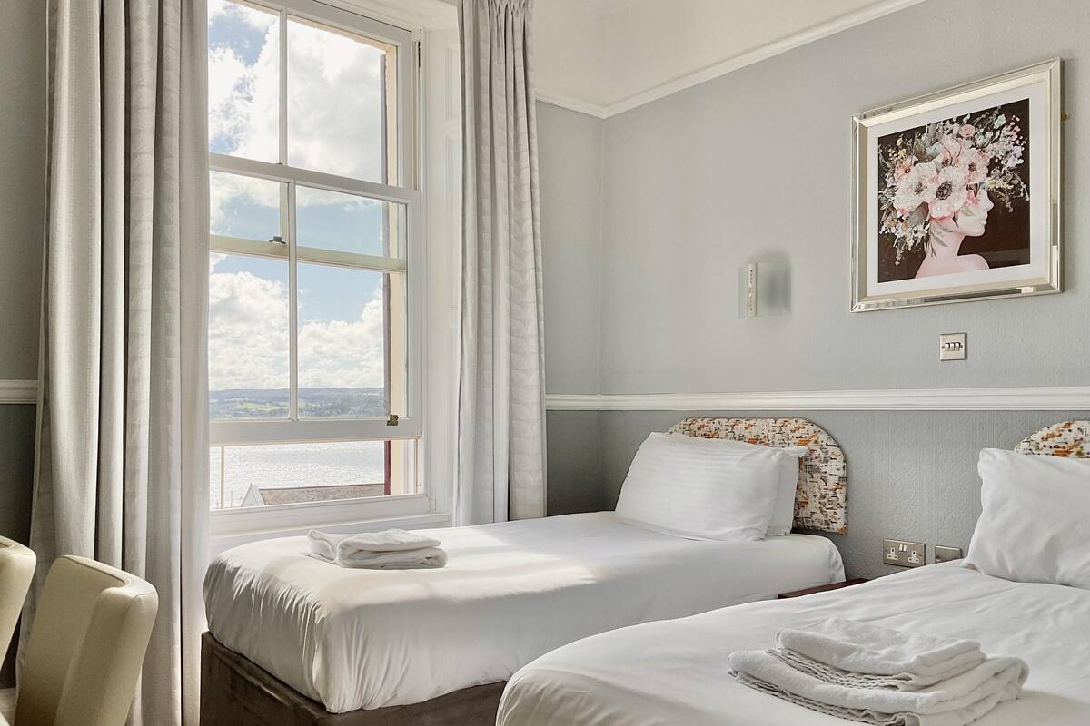 Ett hotellrum med två sängar, ett stort fönster med gardiner, ett inramat blomstermotiv på väggen och utsikt över omgivningen.