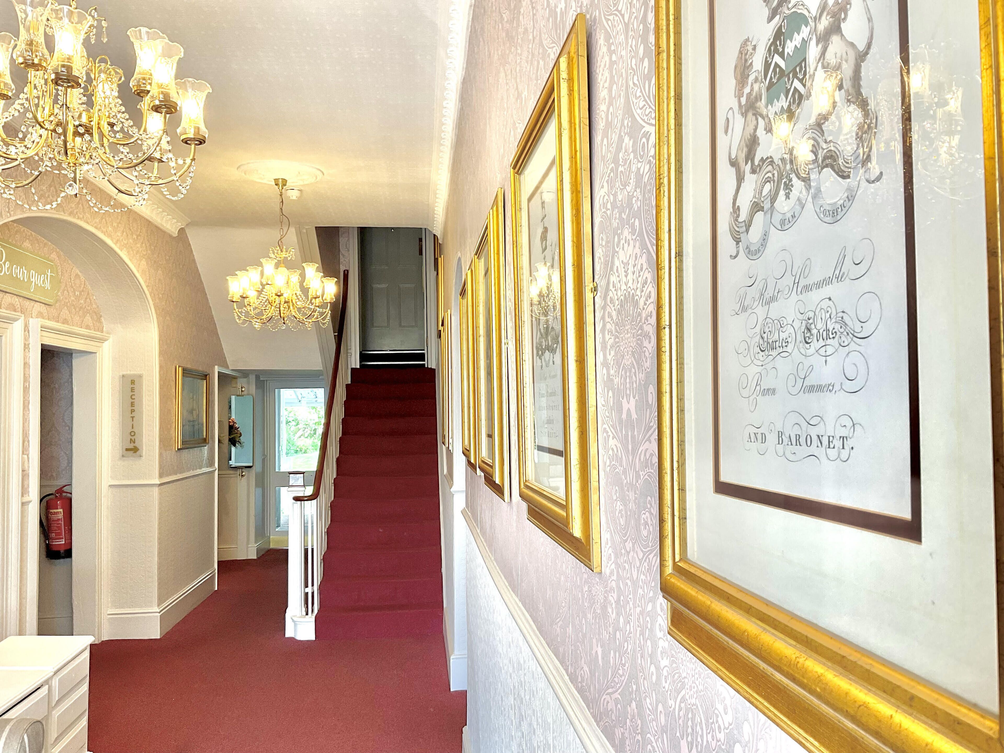 Un vaste hall d’entrée avec un tapis rouge, des tableaux richement décorés dans des cadres dorés, un lustre et une pancarte sur laquelle on peut lire : « Soyez nos invités ».