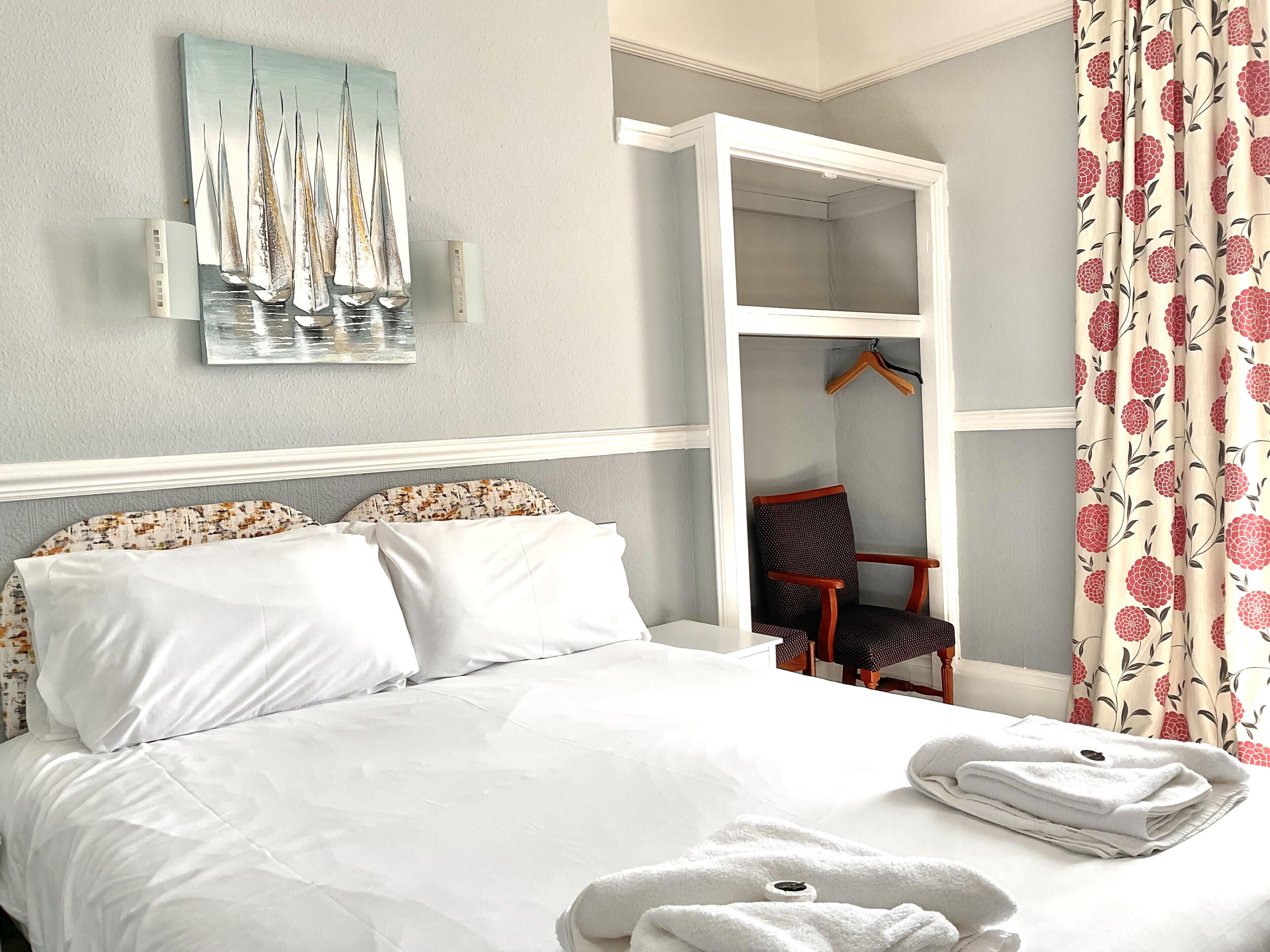 A neatly made bed with white linens, a chair with a floral pattern, and a painting of sailboats on the wall.