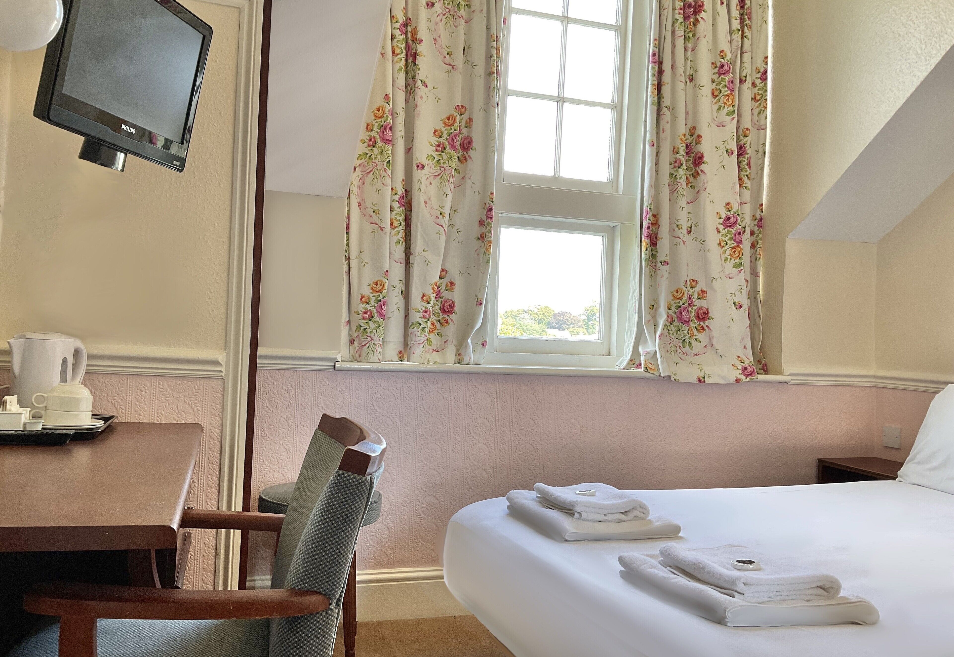 Une chambre d’hôtel comprenant un lit, un bureau avec une chaise, une télévision et une fenêtre agrémentée de rideaux à motifs floraux.